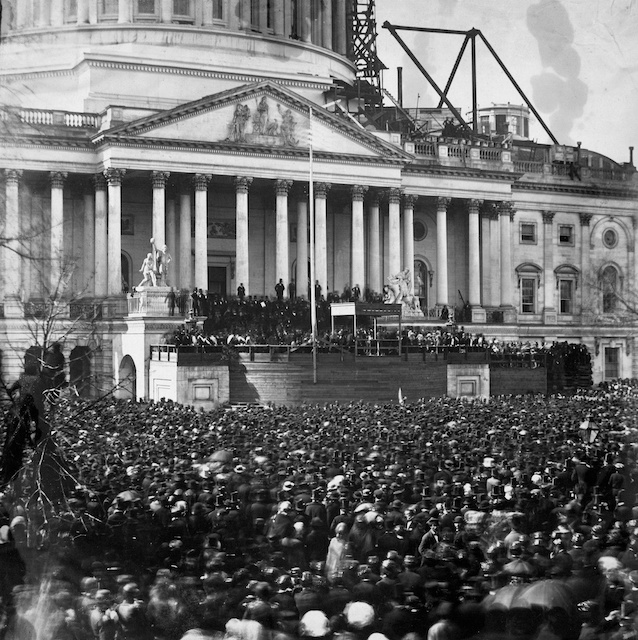 participants and crowd at the first inauguration of President Abraham Lincoln, at the U.S. Capitol, Washington, D.C. Lincoln is standing under the wood canopy, at the front, midway between the left and center posts. His face is in shadow but the white shirt front is visible.  (Wikipedia/Library of Congress).