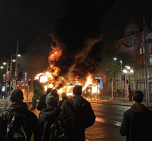 Dubliners watch as a Dublin Bus is engulfed in flames during the 2023 Dublin riot. Photograph taken from O'Connell Bridge, facing northwards towards O'Connell Street. Photo Wikipedia